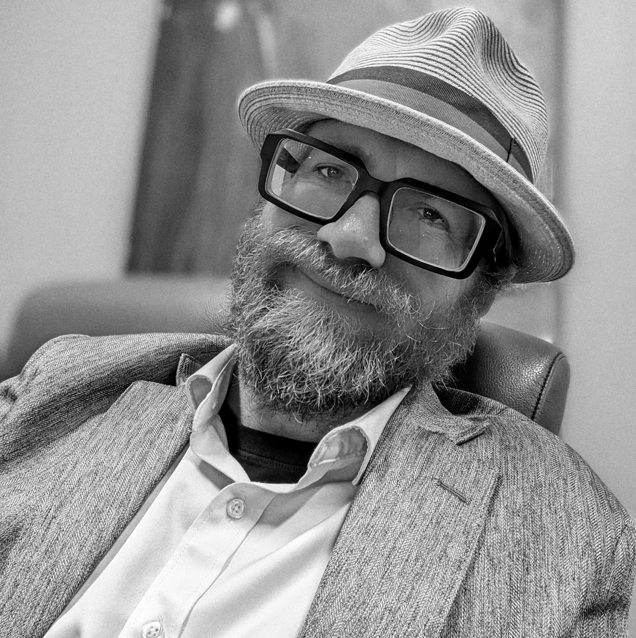 Man with a beard wearing glasses and a striped fedora hat, smiling and seated in an office setting.
