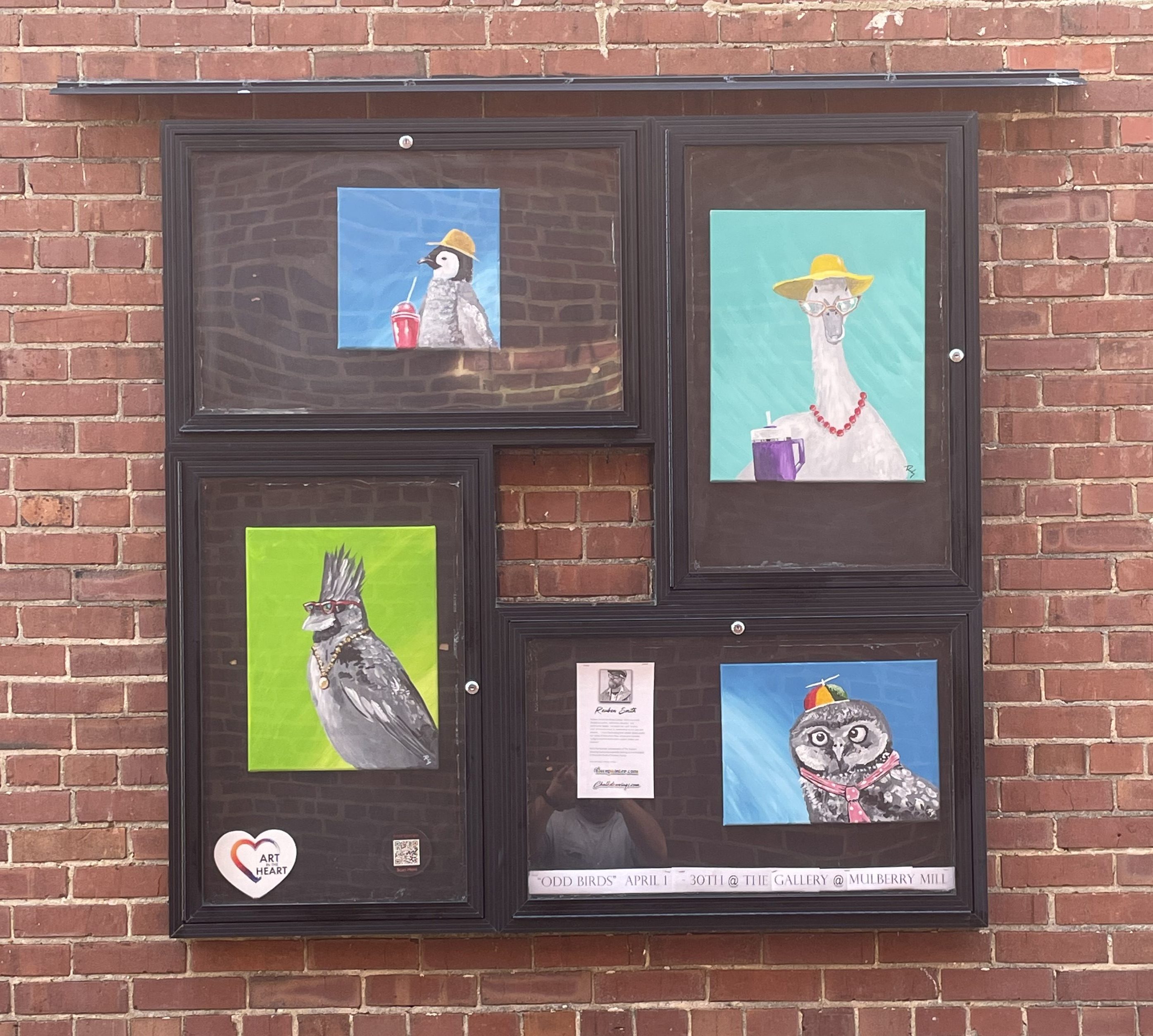 A collection of colorful animal portraits displayed in black frames on a brick wall. The images include a bird with a drink, a llama wearing sunglasses and a hat, a stylish bird with a mohawk and jewelry, and an owl wearing a party hat. Below, there's a poster for an art event titled 'Odd Birds' with details of the gallery.
