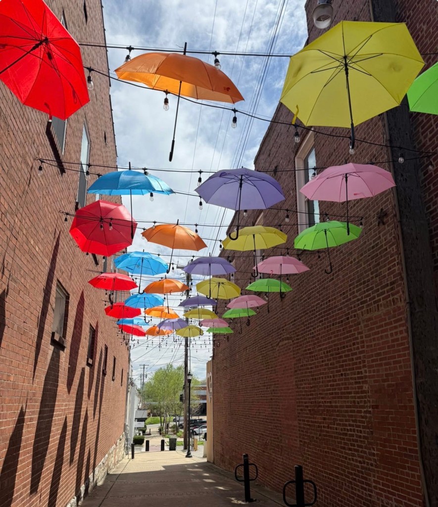 A colorful display of umbrellas hanging above a narrow alleyway, with brick walls and string lights, creating a vibrant atmosphere.